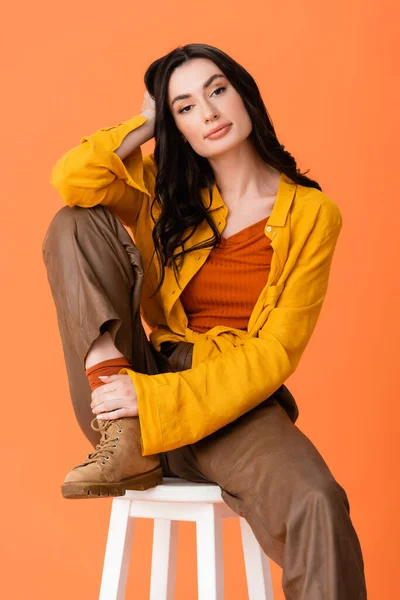 Модна жінка в осінньому вбранні сидить на білому табуреті і дивиться на камеру ізольовано на помаранчевому — стокове фото