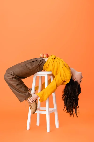 Trendy woman in autumn outfit with apples on belly lying on white stool and touching boots isolated on orange — Stock Photo