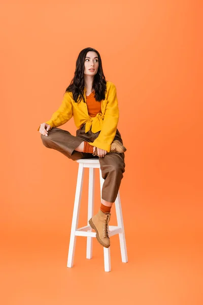 Mujer de moda en traje de otoño sentado en el taburete blanco y mirando hacia otro lado en naranja - foto de stock