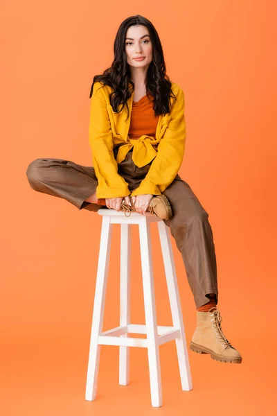 Full length of fashionable woman in autumn outfit sitting on white stool and looking at camera on orange — Stock Photo
