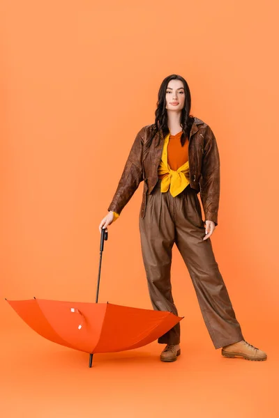 Toute la longueur de la femme à la mode en tenue d'automne et bottes debout avec parapluie sur orange — Photo de stock