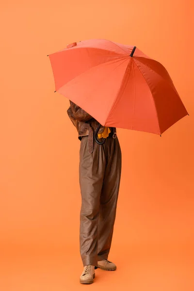 Longitud completa de la mujer de moda en traje de otoño y botas que cubren la cara con paraguas en naranja - foto de stock