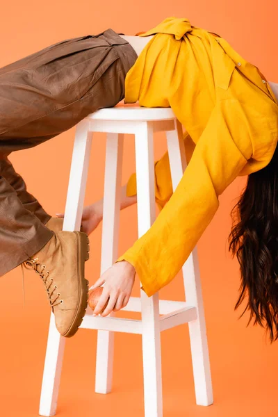 Vista ritagliata della donna alla moda in abito autunno sdraiato su sgabello bianco e stivali toccanti su arancione — Foto stock