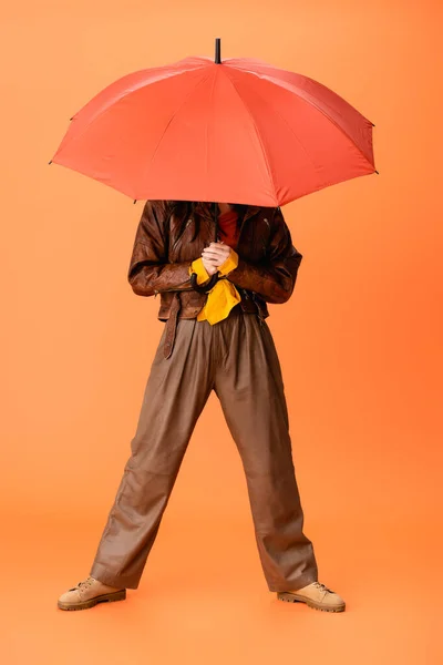 Full length of trendy woman in autumn outfit and boots covering face with umbrella on orange — Stock Photo