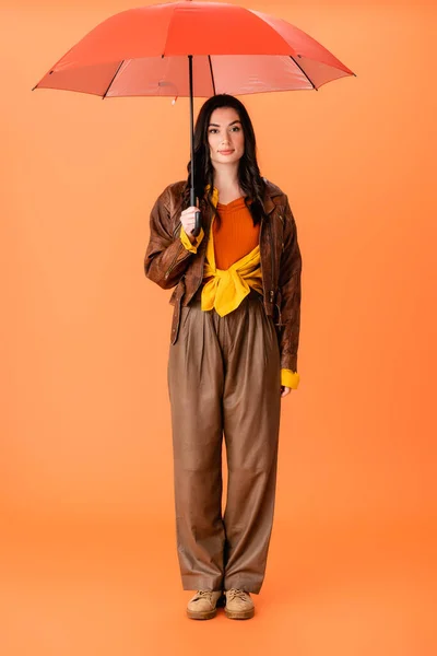 Повна довжина стильної молодої жінки в осінньому вбранні і чоботях, що стоять з парасолькою на помаранчевому — стокове фото