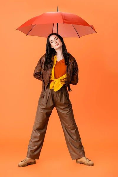 Longitud completa de la mujer joven de moda en traje de otoño y botas de pie con paraguas en naranja - foto de stock