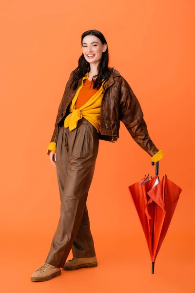 Повна довжина жінки в осінньому вбранні і чоботях, що стоять з парасолькою на помаранчевому — стокове фото