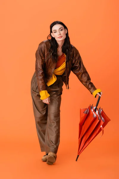 Full length of trendy young woman in autumn outfit and boots standing with umbrella on orange — Stock Photo