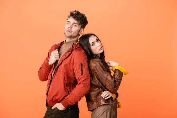 Pareja de moda en chaquetas de cuero mirando a la cámara mientras posan aislados en naranja - foto de stock