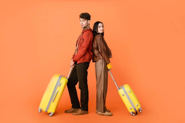 Longitud completa de la pareja de moda en traje de otoño de pie espalda con espalda con equipaje amarillo en naranja - foto de stock
