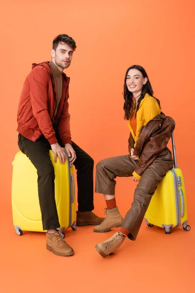 Pareja alegre y de moda en traje de otoño sentado en el equipaje amarillo en naranja - foto de stock