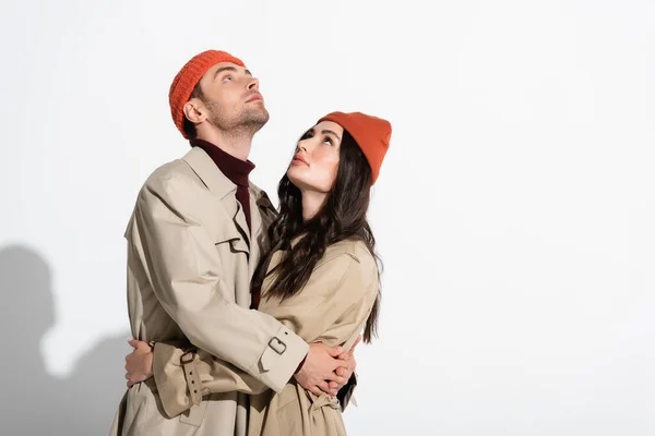 Couple à la mode en bonnet chapeaux et trench manteaux étreignant et regardant vers le haut sur blanc — Photo de stock