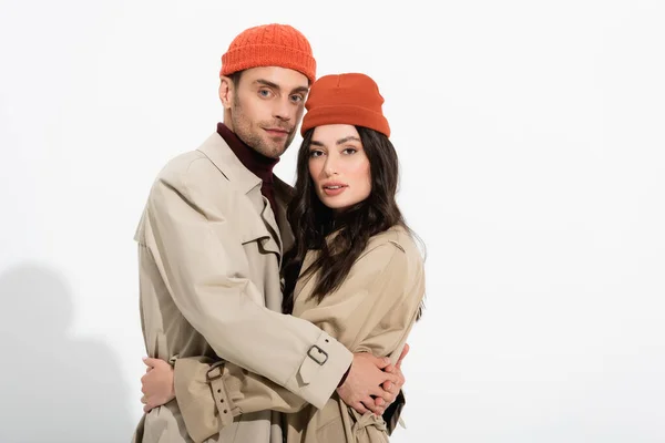 Casal na moda em chapéus de gorro e casacos de trincheira abraçando no branco — Fotografia de Stock