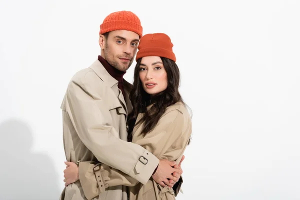 Pareja de moda en gorros sombreros y abrigos de trinchera abrazándose en blanco - foto de stock