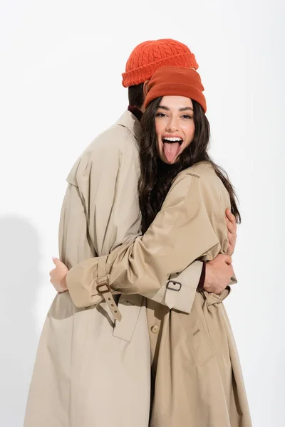 Trendy woman in beanie hat sticking out tongue and hugging man on white — Stock Photo