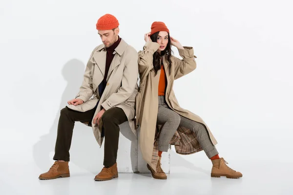 Mujer joven tocando gorro sombrero cerca de hombre de moda sentado en blanco - foto de stock