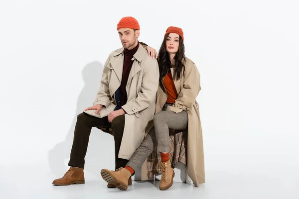 Stylish woman in trench coat sitting near trendy man in beanie hat on white — Stock Photo