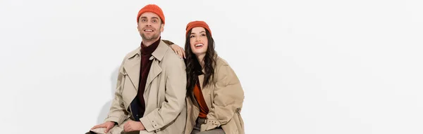 Panoramic shot of stylish woman in trench coat looking up near trendy man in beanie hat on white — Stock Photo