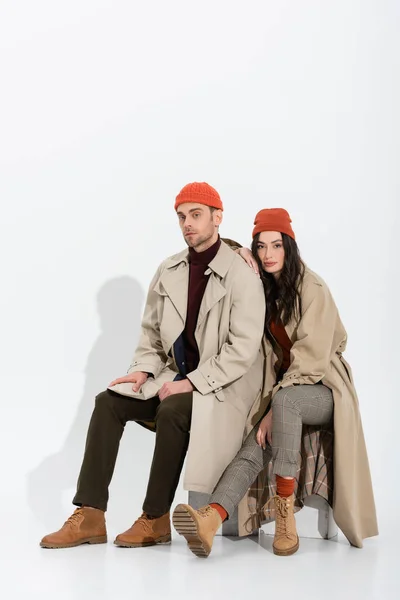 Brunette woman in trench coat sitting near trendy man in beanie hat on white — Stock Photo