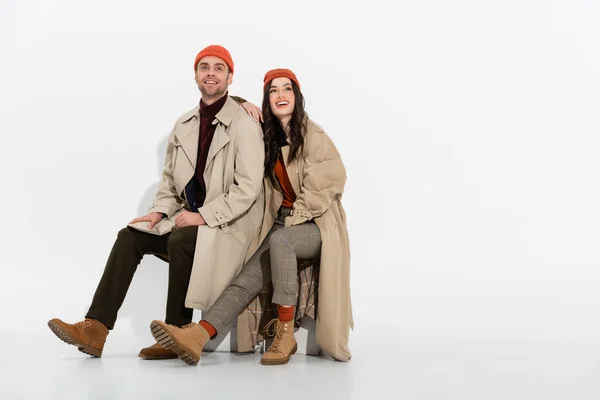 Brunette woman in trench coat looking up while sitting near trendy man in beanie hat on white — Stock Photo