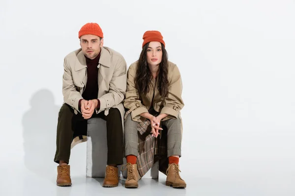 Young brunette woman in trench coat sitting with clenched hands near trendy man in beanie hat on white — Stock Photo