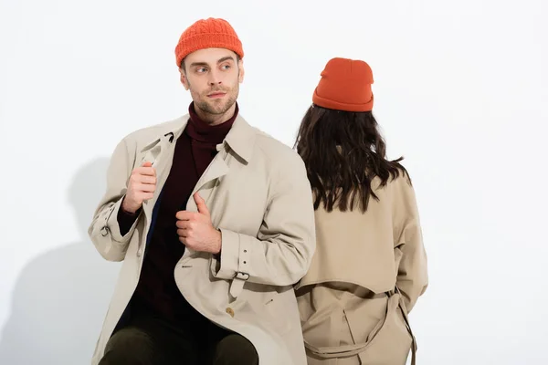 Trendiger Mann mit Beanie-Hut berührt Trenchcoat neben Frau, die auf Weiß sitzt — Stockfoto