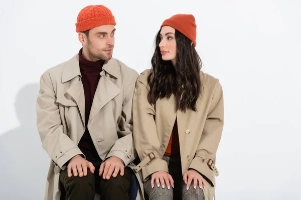 Trendiges Paar in Beanie-Hüten und Trenchcoats sitzt und schaut sich auf weißem Grund an — Stockfoto