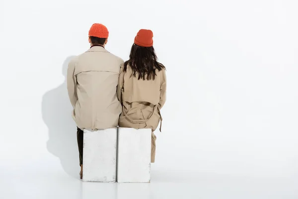 Back view of trendy couple in beanie hats and trench coats sitting on white — Stock Photo