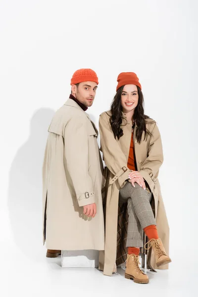 Trendy couple in beanie hats and trench coats looking at camera on white — Stock Photo