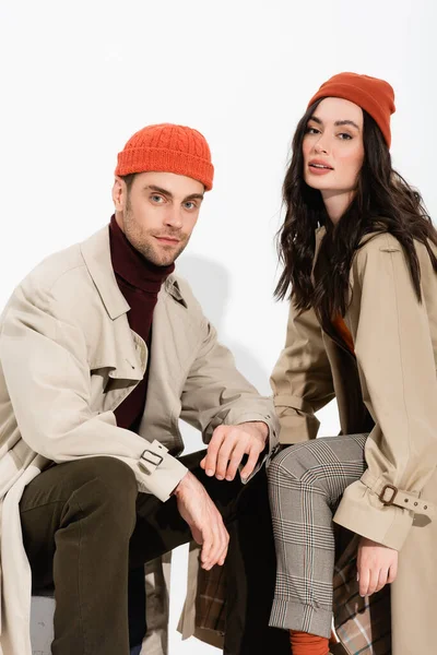 Pareja en gorros y abrigos de trinchera mirando a la cámara en blanco - foto de stock