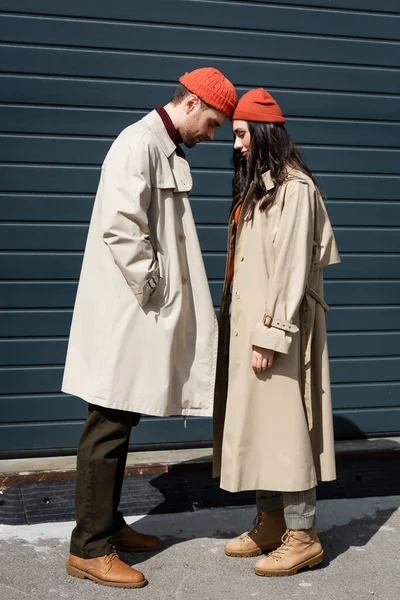 Full length of stylish couple in trench coats and hats leaning on each other outside — Stock Photo