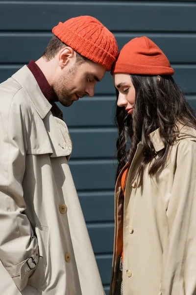 Side view of stylish couple in trench coats and hats leaning on each other outside — Stock Photo