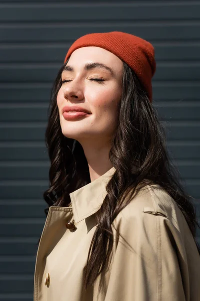 Sunshine on stylish woman with closed eyes in trench coat and beanie hat — Stock Photo
