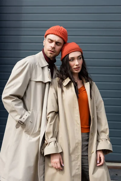 Hombre con estilo en gabardina y sombrero de pie con la mano en el bolsillo cerca de la mujer exterior - foto de stock