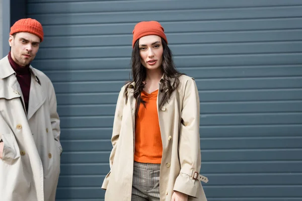 Stylish couple in trench coats and hats standing and looking at camera outside — Stock Photo