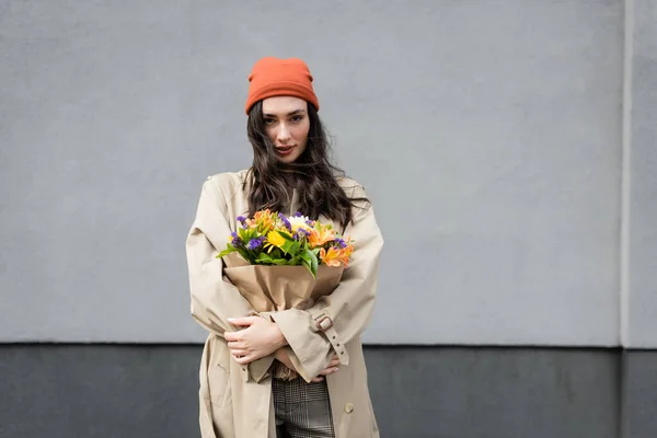 Elegante donna in cappello beanie e trench con fiori vicino alla parete grigia — Foto stock
