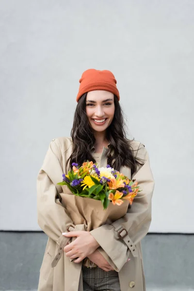 Stylish woman in beanie hat and trench coat holding bouquet of flowers near grey wall — Stock Photo