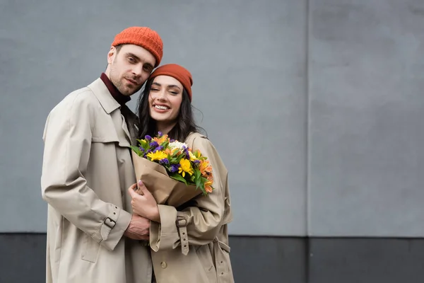 Pareja de moda en abrigos de trinchera y sombreros con ramo de flores - foto de stock