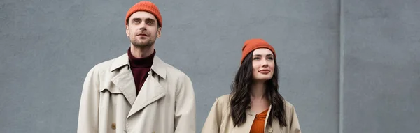 Concepto horizontal de pareja elegante en abrigos de trinchera y sombreros de pie cerca de la pared gris exterior - foto de stock