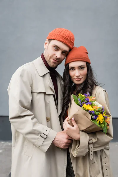 Stylish couple in trench coats and hats holding bouquet of flowers — Stock Photo