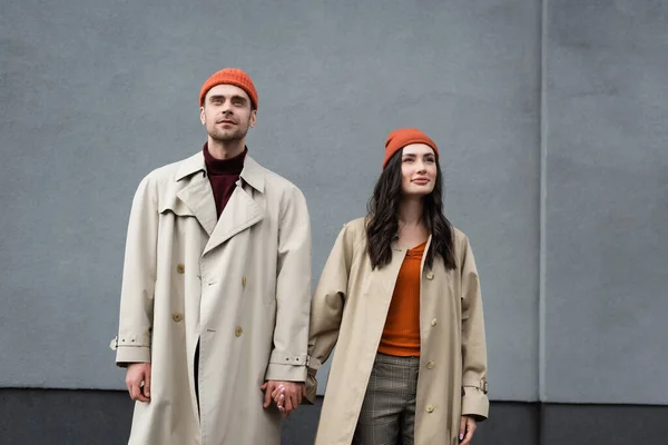 Stylish couple in trench coats and hats standing and holding hands near grey wall outside — Stock Photo