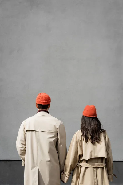 Vista posteriore di coppia in trench e cappelli in piedi vicino alla parete grigia — Foto stock