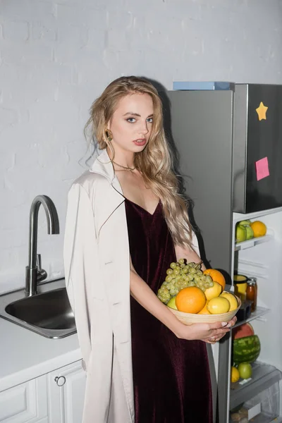 Jeune femme blonde en trench coat et robe de velours regardant la caméra tout en tenant bol avec des fruits frais — Photo de stock