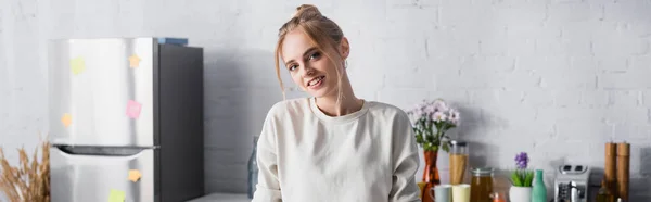 Plano panorámico de la mujer rubia sonriendo a la cámara mientras está de pie en la cocina - foto de stock