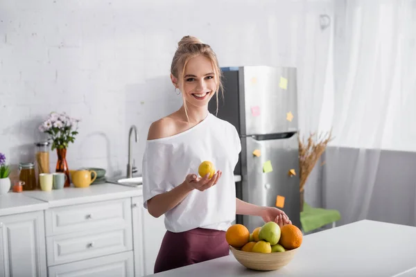 Mujer rubia sonriente sosteniendo limón mientras está de pie cerca de un tazón con frutas frescas - foto de stock