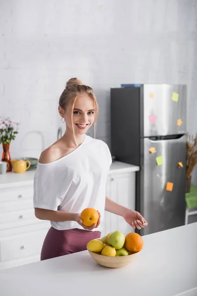 Happy woman in white t-shirt smiling at camera while holding ripe orange near bowl with fresh fruits — Stock Photo