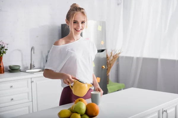 Mujer rubia feliz en camiseta blanca mirando a la cámara mientras vierte té en la cocina - foto de stock