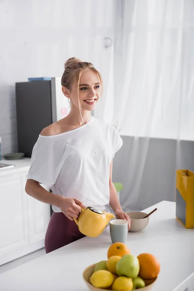 Glückliche Frau im weißen T-Shirt schaut weg, während sie Tee in Tasse gießt — Stockfoto