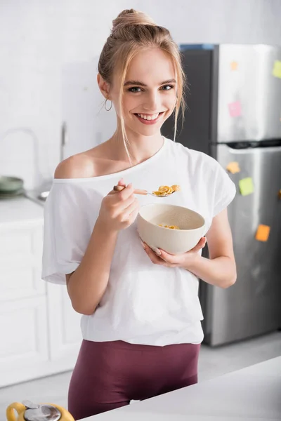 Blonde woman in white t-shirt smiling at camera while eating cornflakes for breakfast — Stock Photo