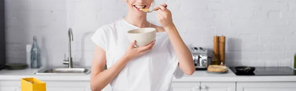 Vue recadrée de femme heureuse en t-shirt blanc mangeant des flocons de maïs pour le petit déjeuner, culture panoramique — Photo de stock